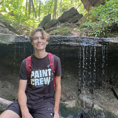 Isaac Abram posing in front of a trickling waterfall cascading off of a rock shelf in the woods.
