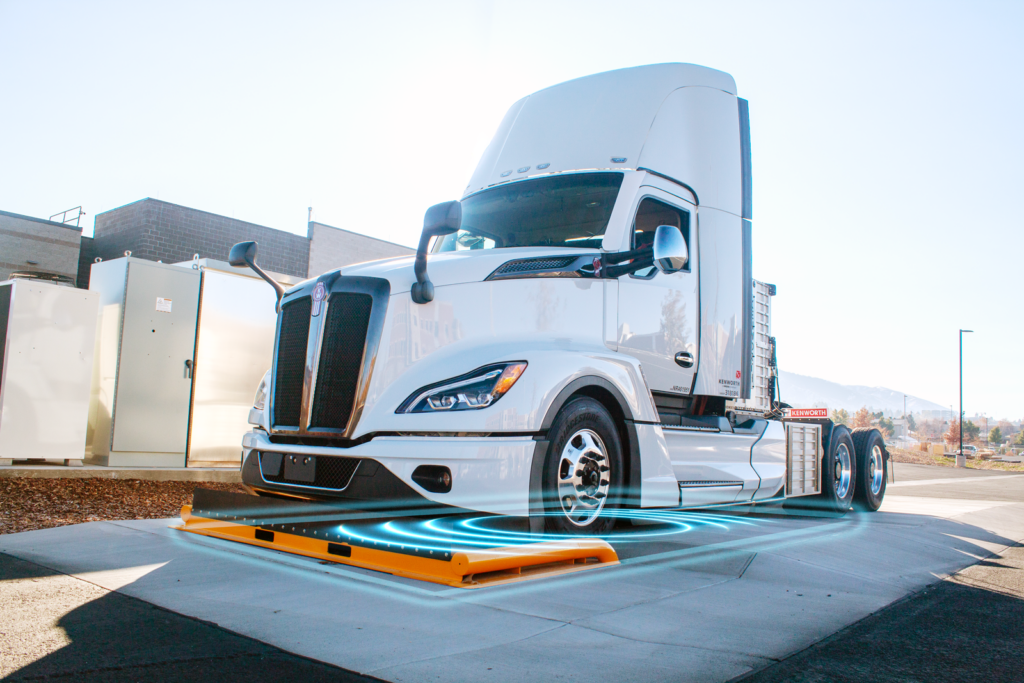 The Falcon — a Class 8 semi truck designed by Kenworth — sits on ASPIRE's 1-megawatt charging pad. Electric blue illustrations demonstrate the wireless power transfer that charges the truck without needing to be plugged in.