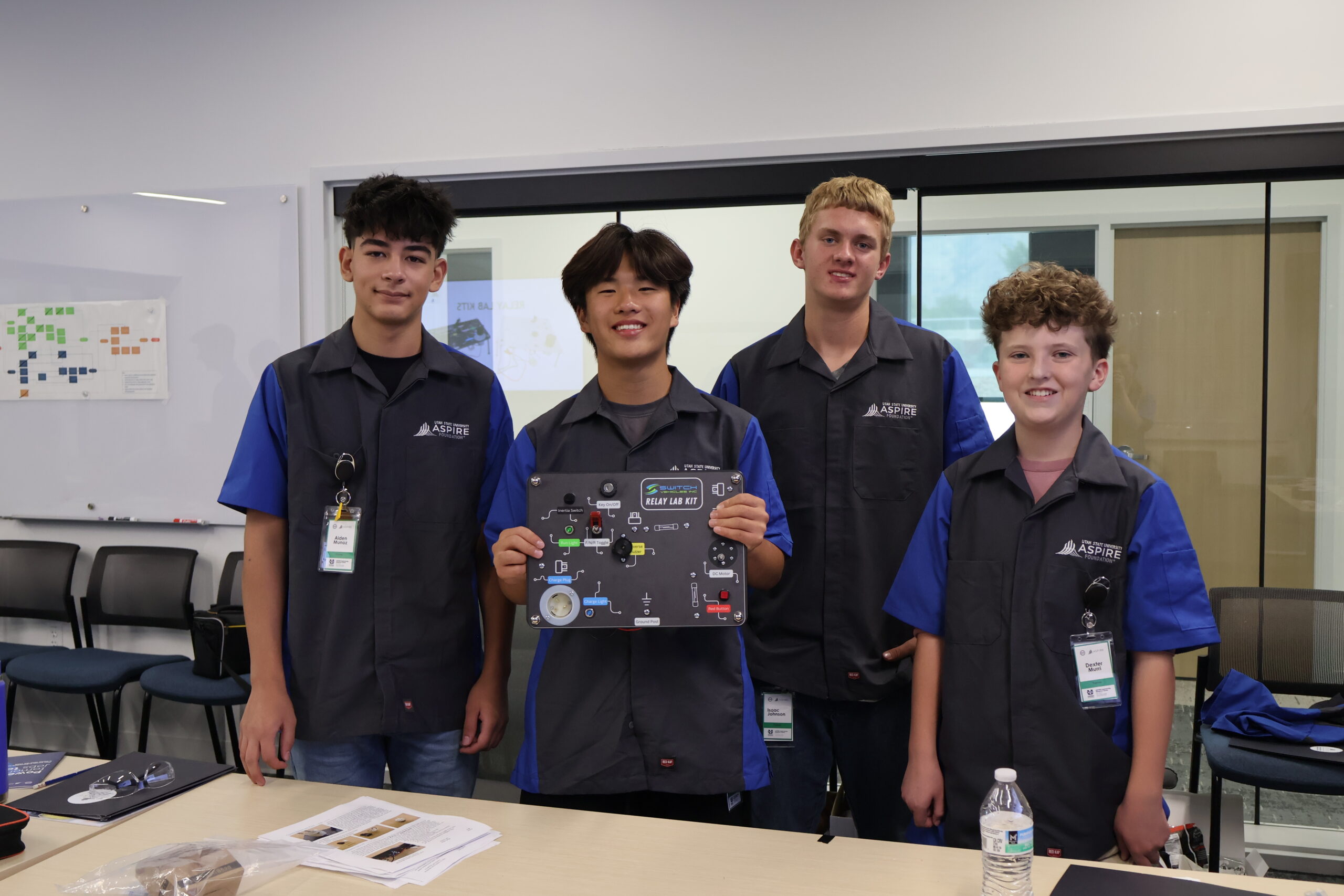 four students wearing gray mechanical shirts with blue sleeves hold up an electric wire board while smiling