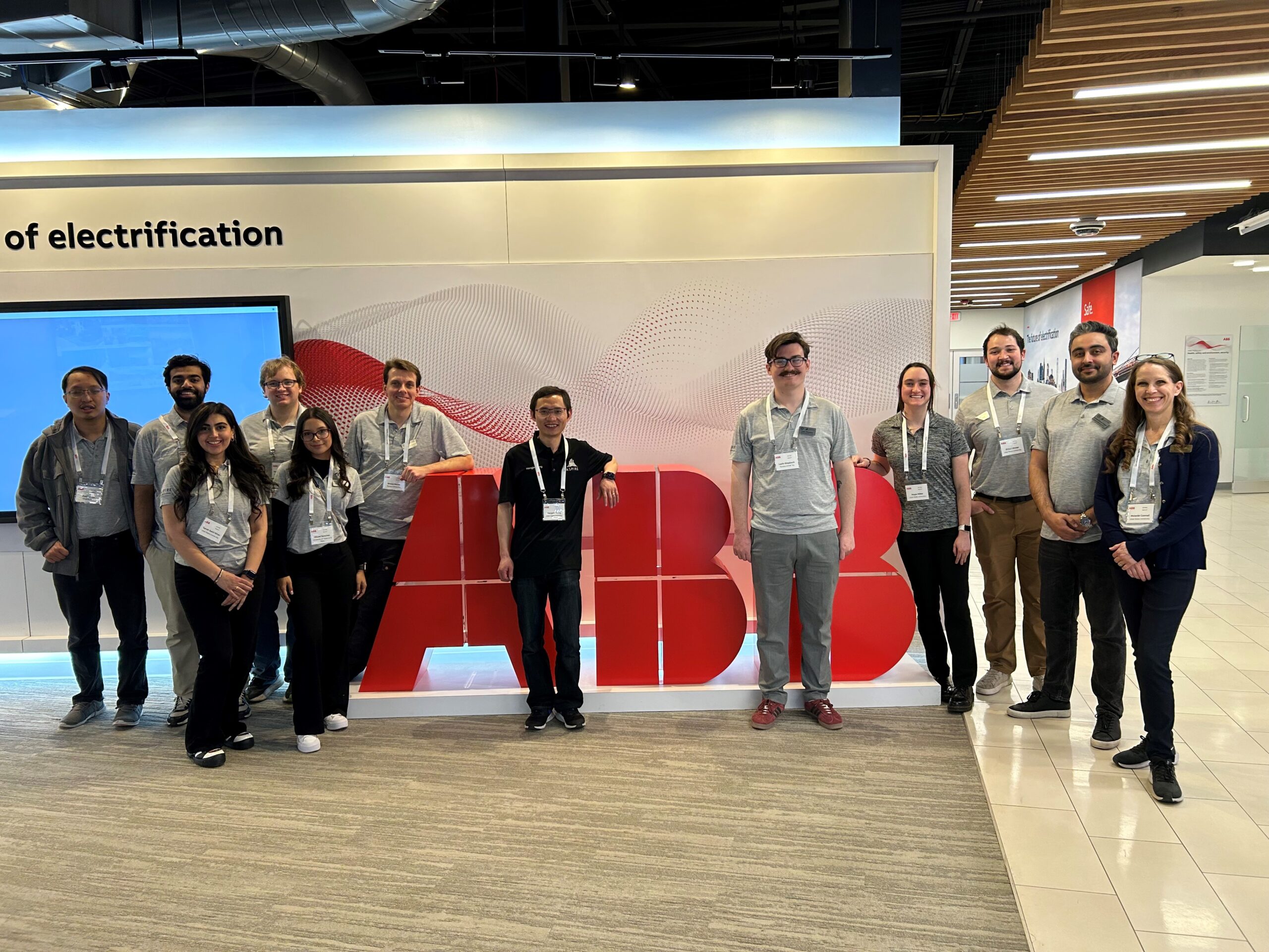 A group of ten students stand inside a building with fluorescent lights shining down the group and offset hallway in the photo. Students stand between large letter blocks, with a student and letter alternating for the "ABB" acronym.