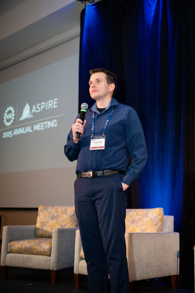 A man wears a blue shirt with blue trousers and a brown belt. He holds a microphone with white marbled couch behind him. Behind the couch, a blue curtain with a light shining the curtain shines up to the ceiling. NSF ASPIRE Annual Meeting is projected on to a screen behind him as he faces toward the audience.