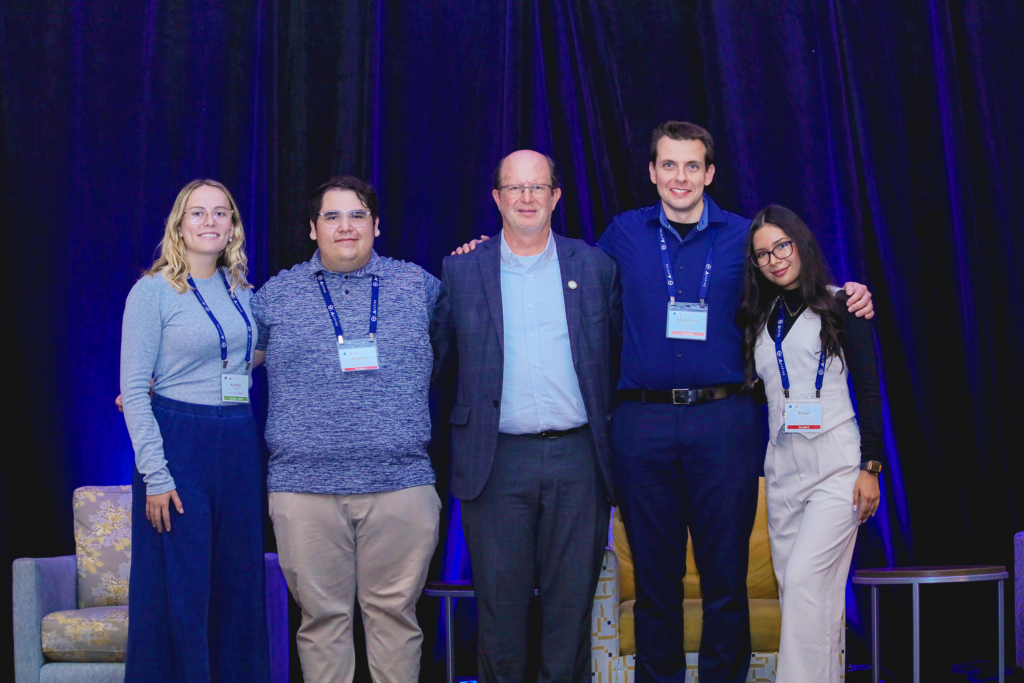 Five people stand in front of a blue curtain with arms surrounding one another. They wear professional attire, and smile at the camera.