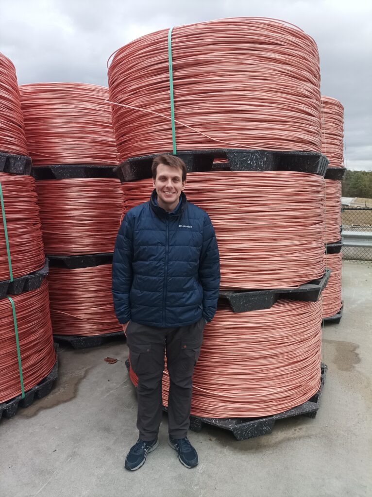 A man in a blue jacket wearing black pants and blue shoes stands in front of industrial sized rolls of copper, three stacked on top of each coil, so they loom over the man.