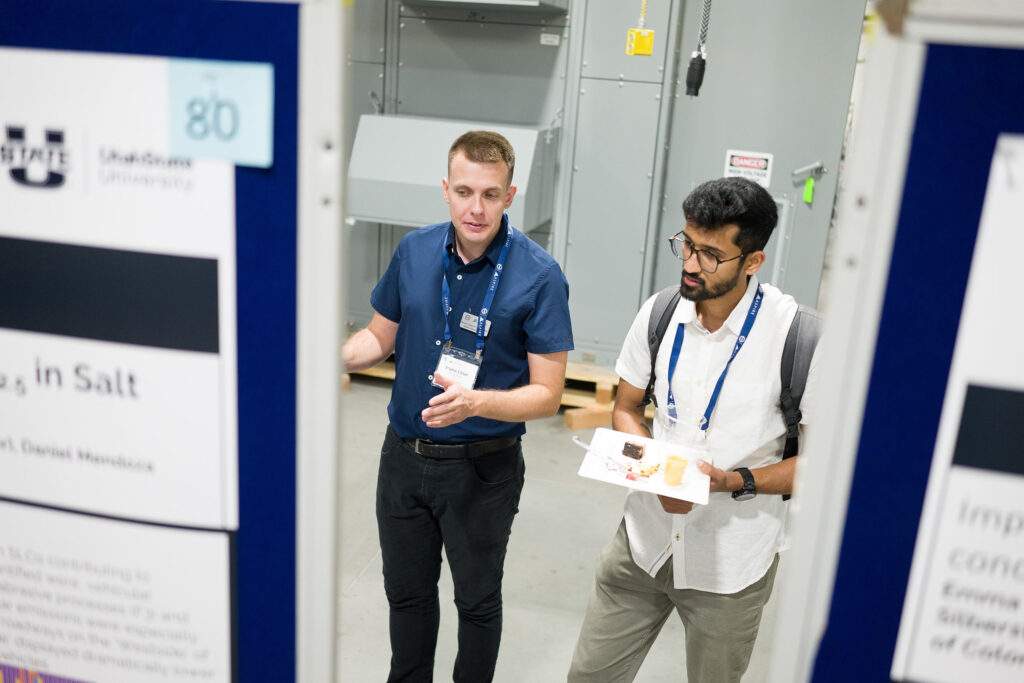 Two men stand in-between two research posters in the center of the photo. The two posters in the image block the left and right thirds of the image. Hands are outstretched in explanation while their eyesight is facing the research poster. The camera angle, we cannot see what they are viewing as they face the camera.