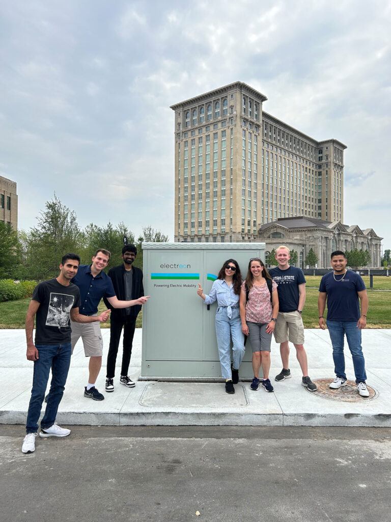A group of students stand next to and point to an Electreon Power box. In the background there is a line of trees and a large multi-storied building with a cloudy gray sky with some blue coming through.