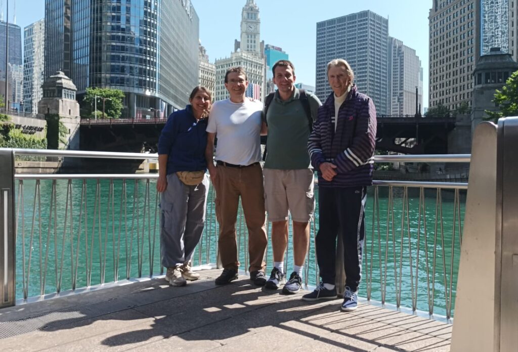 Four people stand with their backs to a pier overlooking a river with city skyscrapers in the background. The sky is a clear blue with glass buildings reflecting black the blue sky and grey buildings. The water is a blue green surrounding the background of the pier to the people's hips. The posed individuals smile to the camera standing on a cement and metal designed outlook.