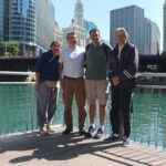 Four people stand with their backs to a pier overlooking a river with city skyscrapers in the background. The sky is a clear blue with glass buildings reflecting black the blue sky and grey buildings. The water is a blue green surrounding the background of the pier to the people's hips. The posed individuals smile to the camera standing on a cement and metal designed outlook.