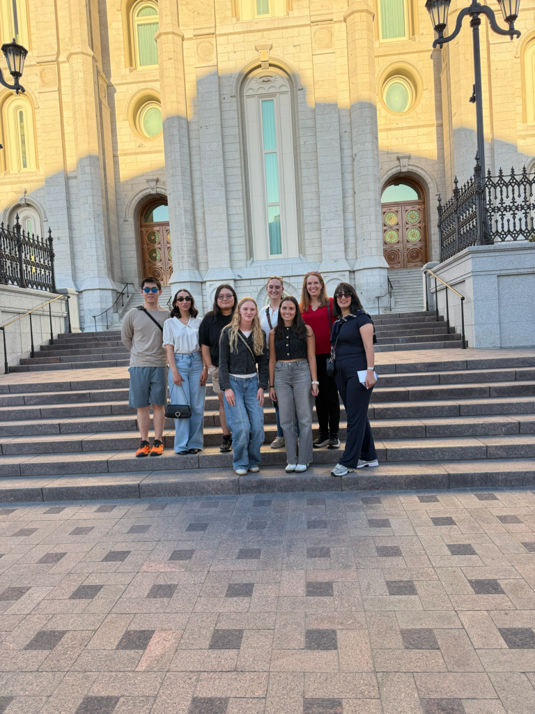 A group of eight people stand of the steps of a grey and cream concrete entry way building. The whole group smiles at the camera. A shadow overcasts the group onto the Romanesque and medieval inspired architectural building behind the group. Behind the group there are two sets of large double doors, a copper amber with round golden decal on it.