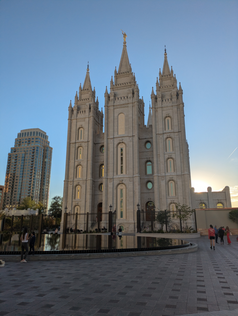A large building with gothic revival and Romanesque architectural influences takes up a large portion of the photo. The temple shown in the picture has three main spires with a grey-cream brick to it, and gold accents and clear blue glass panes in the front of the building. In front of the large temple building, there is a reflection pool and concrete walkway outside in a city area. The sun is setting over part of the temple building and the sky is blue, with the bottom edge of the sky showing, orange. Small groups of people walk along the concrete walkways, and a large skyscraper is seen left of the temple building in the background, with trees and a black metal fence lining the edge of the temple walkway.