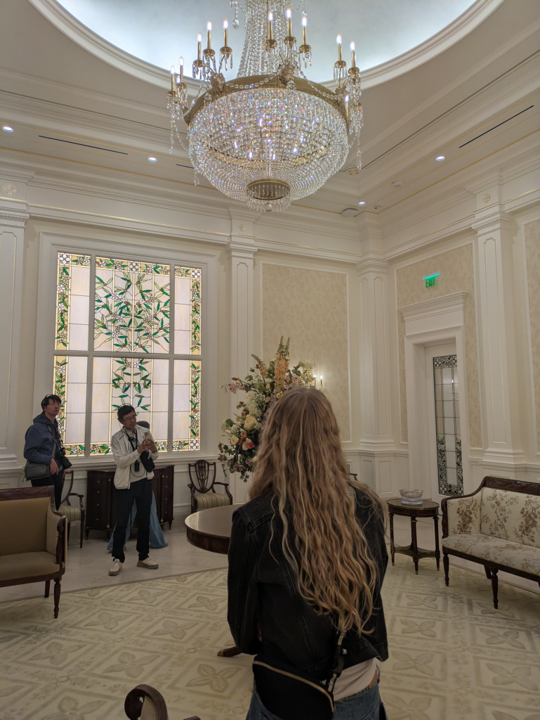 There is a group of four people standing and looking at the walls, and ceilings of a cream and white room. A woman in center frame looks up at a chandelier- decorated in many crystals with light refracting into small rainbows. Behind the woman there is a large stained-glass window with cream, green and whites making floral and green decorations.