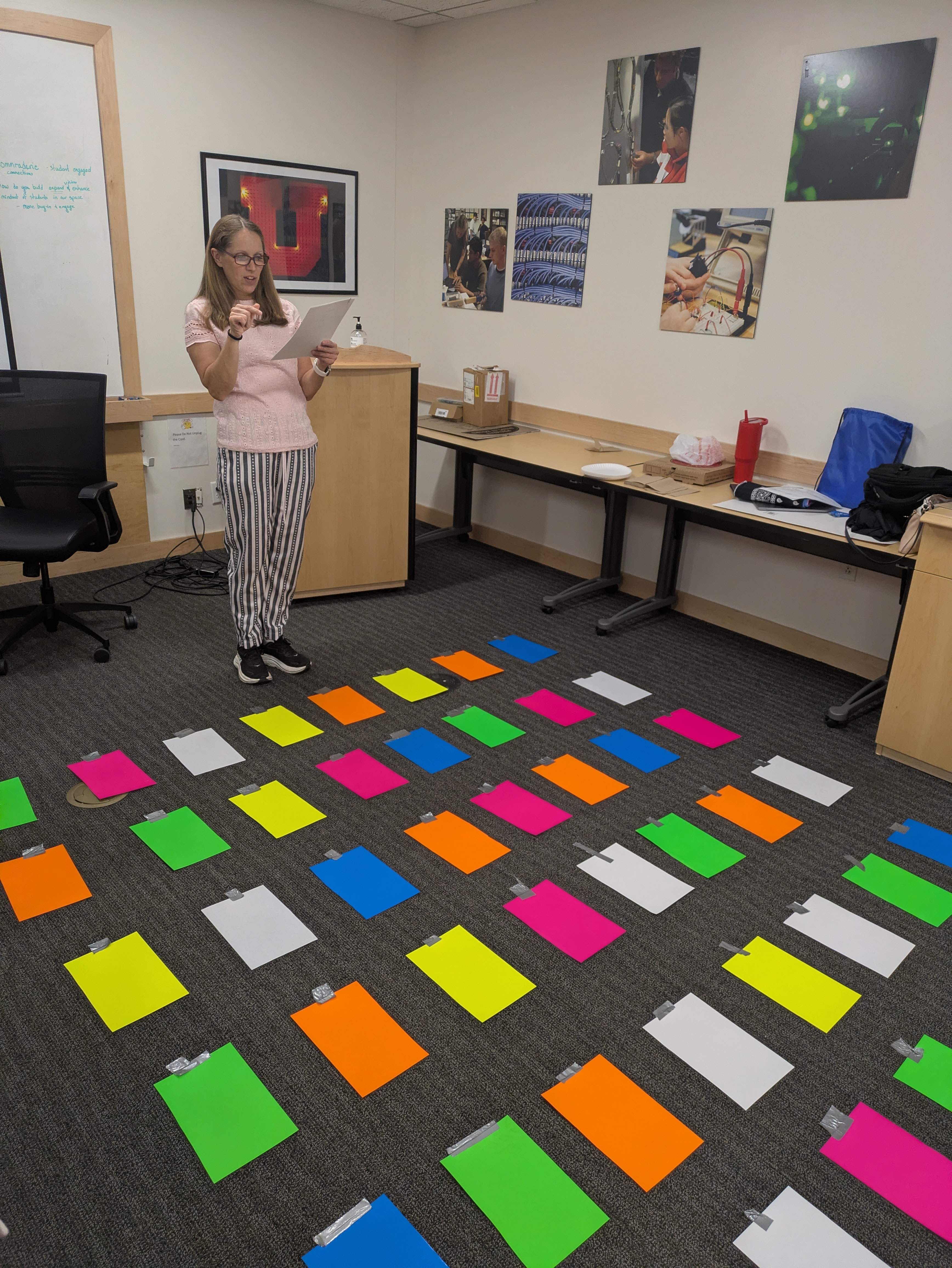 A woman stands at the front of a classroom facing a large array of monochrome colorful sheets of paper taped to the ground individually in columns and rows. The color of paper on the ground is white, yellow, pink, green, and blue. The woman stands holding a piece of white paper in one hand, with her other hand articulating what she is explaining. She wears glasses and her eyes are focused on the white sheet of paper she is holding. There are square pictures of electrical cables, wiring, people interacting with one another, and the University of Utah "U" photo behind the speaker. Student Leadership Retreat 2026 SLC. Melanie Conrad