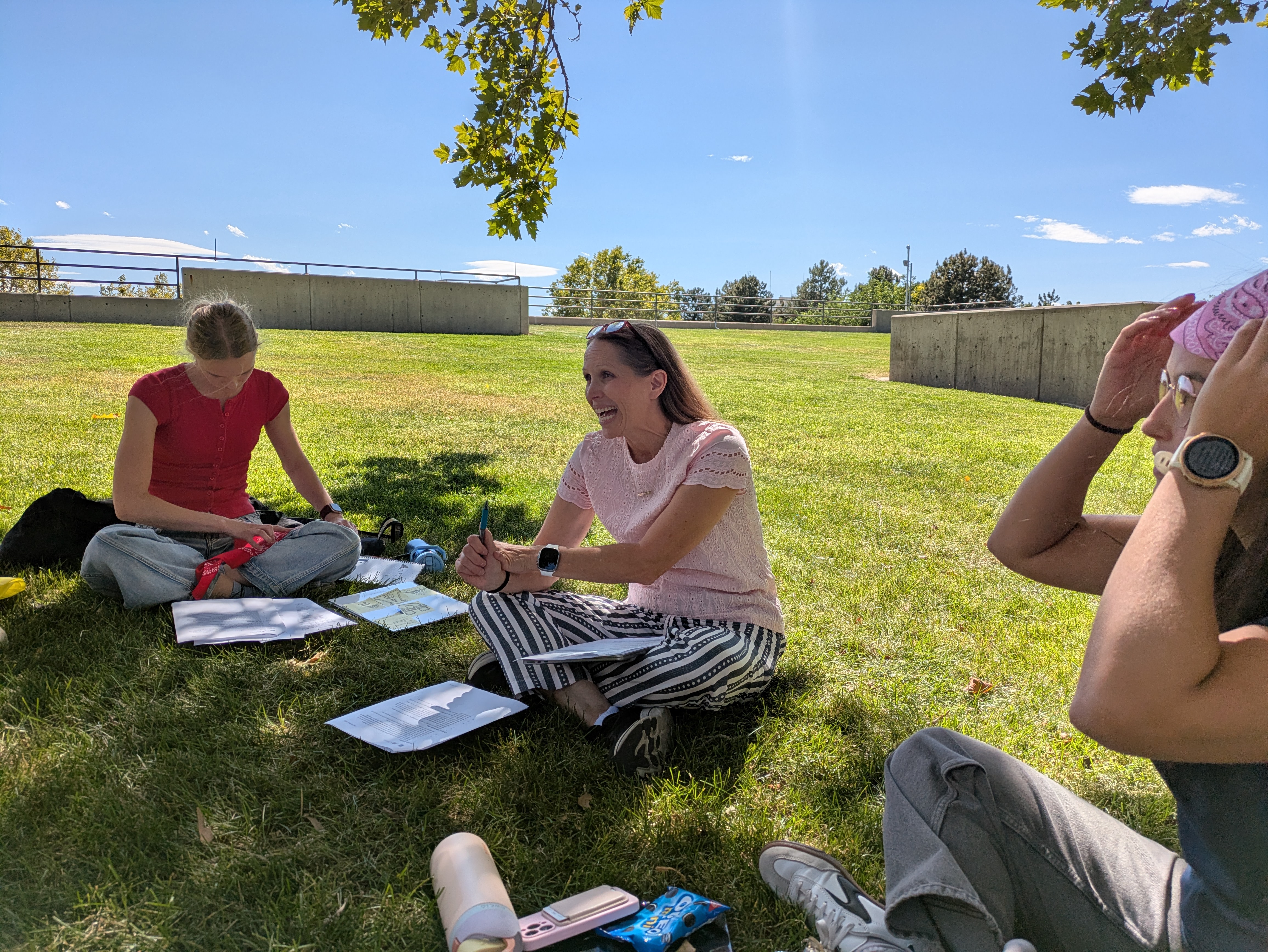Three people sit on a green grassy field that takes up the 65% of the background, with the other 35% being a blue sky with very few clouds in it. The group of people sit in shade a tree is providing while sitting sit-cross-applesauce. The woman in the middle of the three people is laughing looking at someone off camera. Another woman sits to the left of the laughing woman wearing a red shirt, while looking at multiple packets overlaying each other on the ground. The woman on the right of the laughing women wears a pink bandana, with her hands seemingly adjusting the bandana she is wearing on her head.