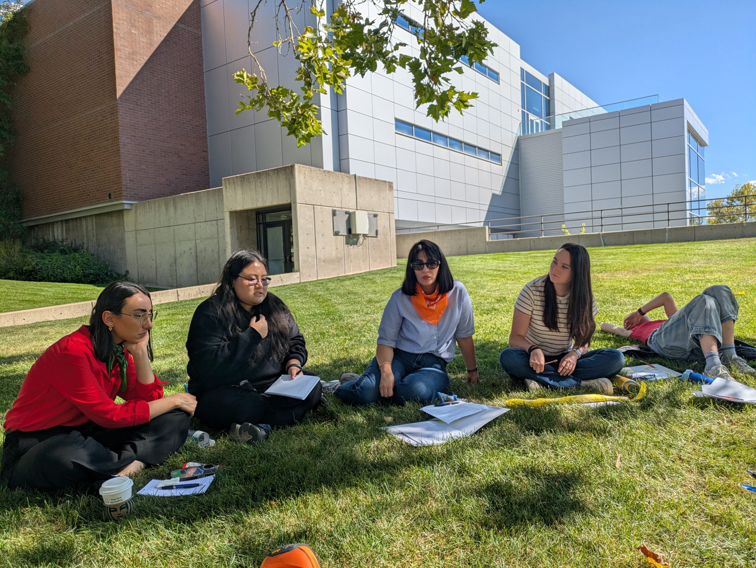 A group of five women sit on a green grass lawn next to a grey concrete and red brick building, with a blue sky in the background and a tree branch with green leaves coming into view from the center of the photo. Three women wear long sleeved shirts with one of the women's sleeves rolled up to her elbows. The other two women wear short sleeved shirts, and one of them is laying down. They are discussing and listening to one f the members of the group speak (the speaker holding a notebook and packet while speaking as the other women listen).