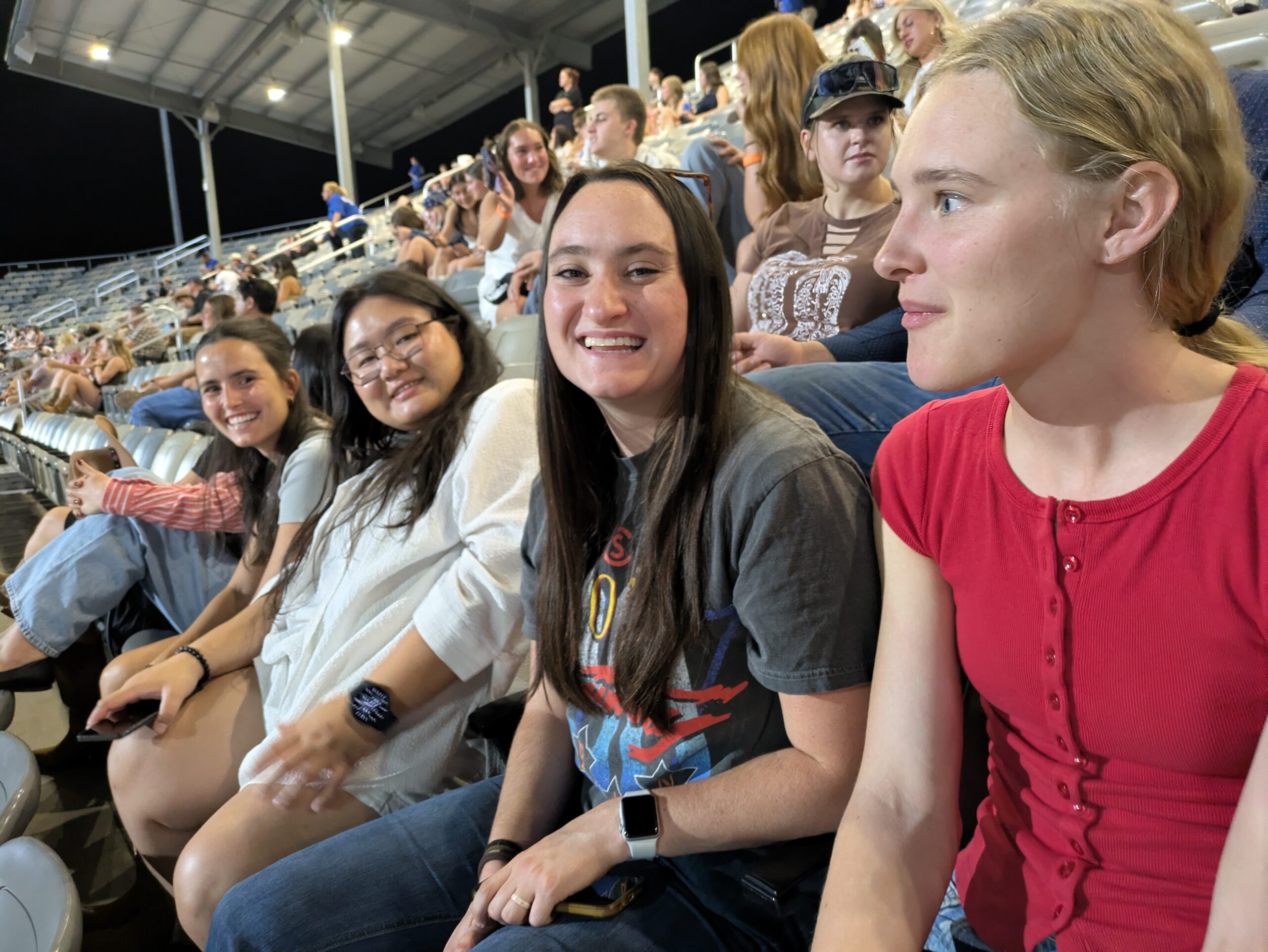 Four people sit next to one another in a large outdoor stadium with a packed crowd behind the row of people smiling toward the camera. The sky showing in the picture is an ombre from orange to a darker blue; the sun is setting.