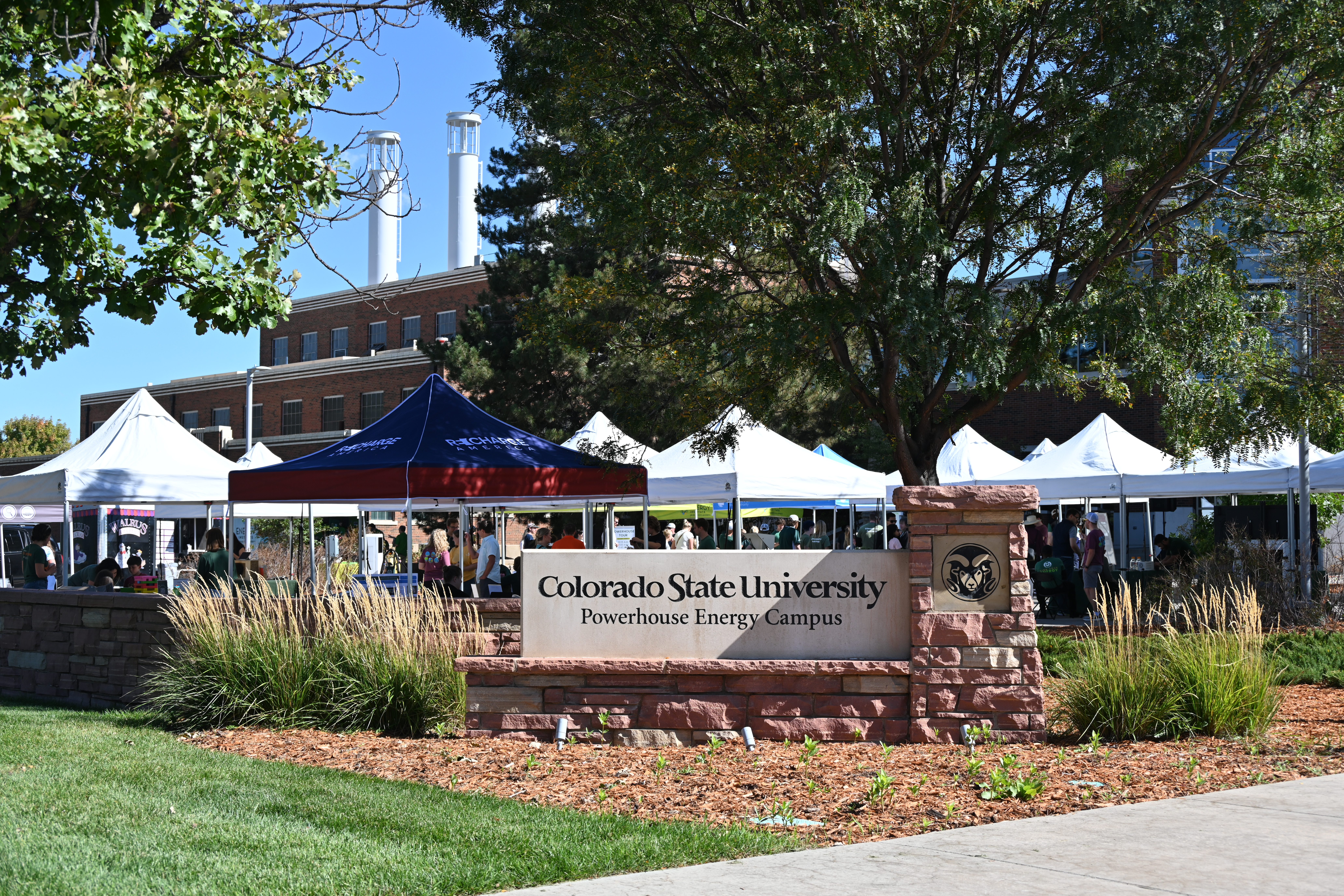 A large outside area at Colorado State University Powerhouse Energy Campus with a factory-like building in the background, has a large amount of pop up tents and booths behind the Colorado State University Powerhouse Energy Campus concrete stone wall sign.