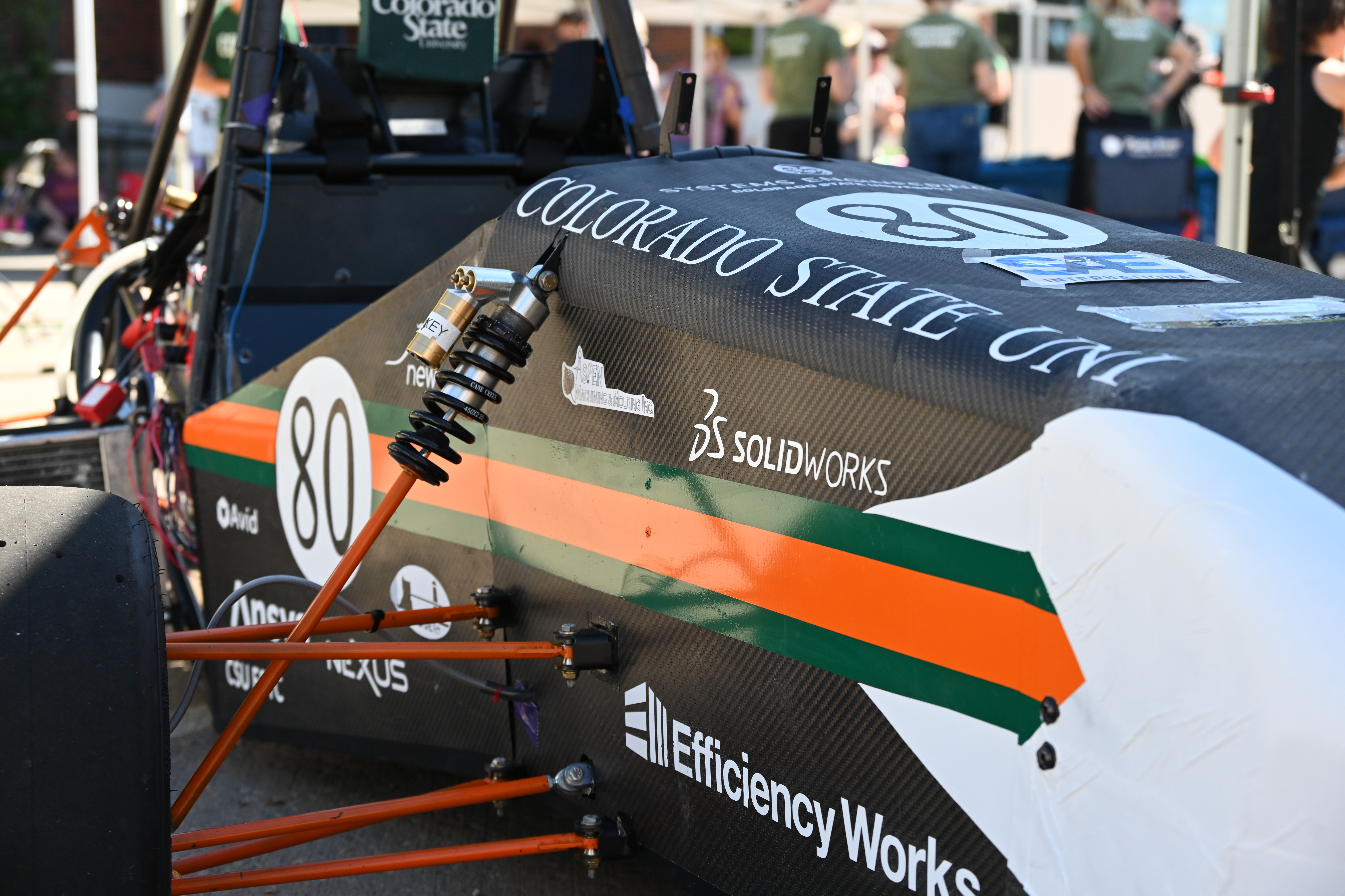 A close up of a race car with Colorado State Uni logo on it, with orange and green stripes down the edge of the car with the black number 80- painted on the circle with a various amount of logos: SolidWorks, Efficiency Works, Arid, Lexus, etc. Orange wires holding out the tire of the racecar are in close focus, with green t-shirts in the background.