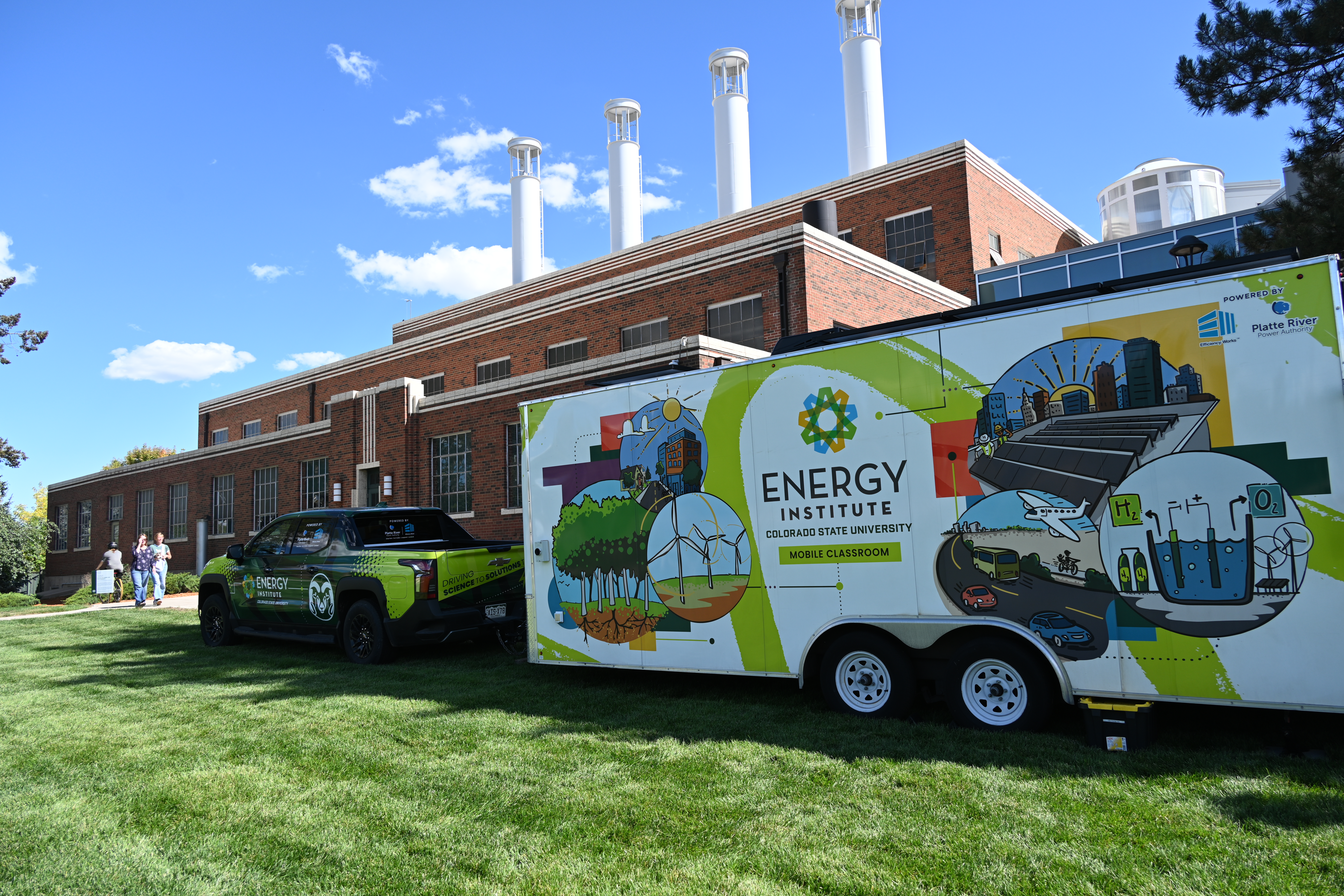 A green lawn with a green truck with the Colorado State Logo on it hauling a Energy Institute trailer with renewable energy options: solar panels, wind turbines, chemical plants, etc. decorate the trailer wrap. Behind the green truck-trailer and lawn is a red brick factory like building with Vertical axis wind turbines replaced the original smoke stacks of the building.