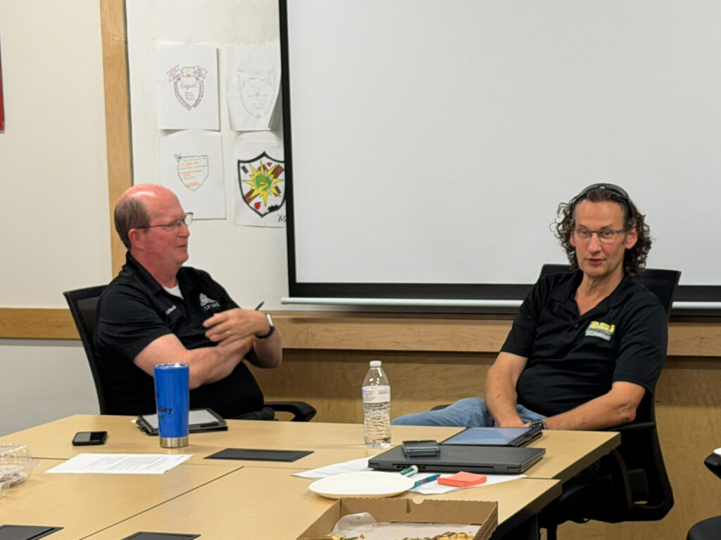 Two men sit at a conference table. A large white board is behind them, and they are seated relaxed in the conference room. They both wear black shirts with one of the men balding, and the other with chin length hair.