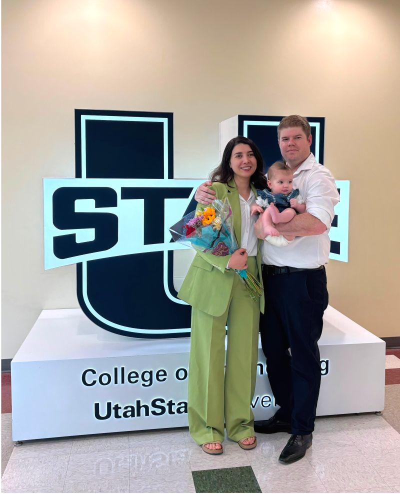 A woman in a green pantsuit and a man stand close together, the man holding a baby in his arms, and the woman holding a bouquet of flowers in front of a Utah State University glowing structure celebrating graduation.