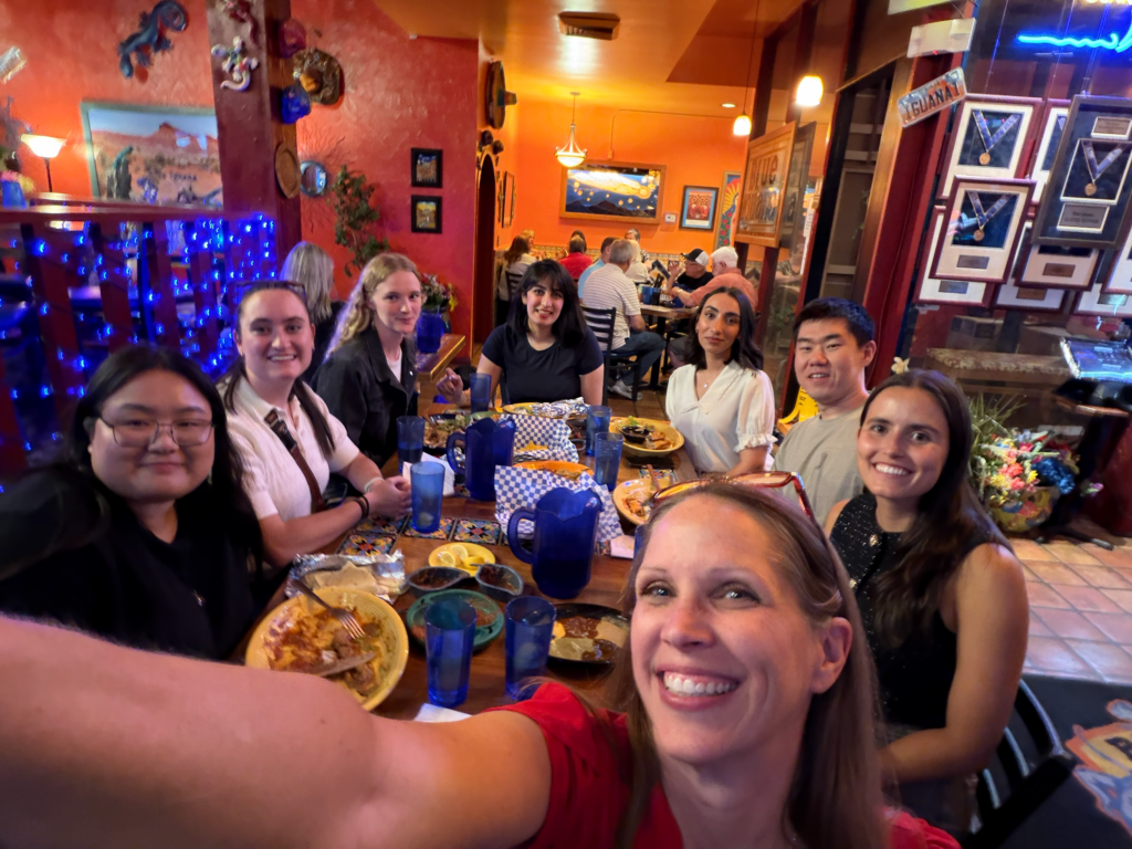 Participants in the ASPIRE Student Leadership Retreat pose for a group photo during dinner at a restaurant, seated around a large table with meals, drinks, and colorful décor in the background.