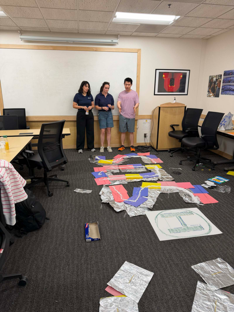A group of three people in a University of Utah small conference room, look at the floor discussing a strategic plan for the team building exercise they are part of. Aluminum foil sheets and multicolored sheets of paper are arranged in an "SOS" shape. A conference room table and chairs are pushed to the side of the classroom with a blank white board behind them.
