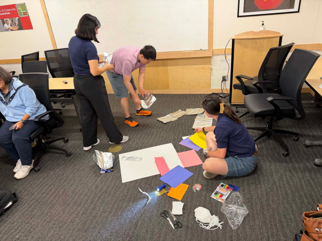 A group of four people in small conference room, stand and kneel over the floor with arms rearranging aluminum foil sheets and multicolored sheets of paper with scissors, markers, and flashlight on the ground. A woman sits in a conference room chair next to the group of students arranging the sheets of paper and tinfoil.