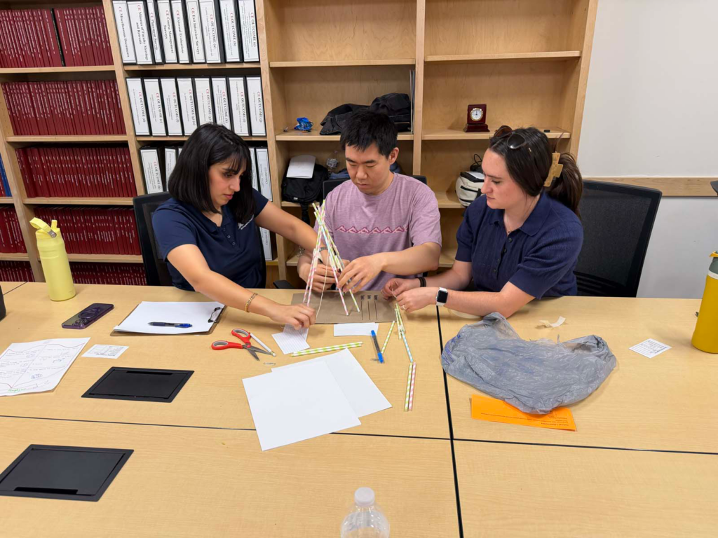A group of three people sit at a conference table with their hands holding a structure together made out of striped paper straws and cardboard. The students are focused on the structure. There are straws, paper, scissors, a plastic bag, a phone, and metal water bottles on the table. Behind the three people is a large built-in bookcase with large red textbooks in one collum, and thicker white three-ring binders in the next collum.