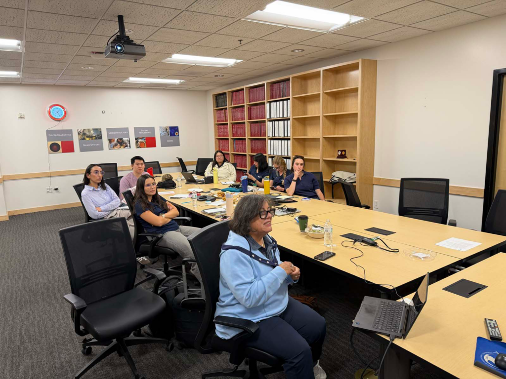 A group of eight people sit around a conference table with laptops open, and food around the table. There is a built-in bookshelf and engineering signs on the back wall with a red clock and a built-in bookcase to the right of the photo. On the ceiling there is a projector with a projecting light, and the group of people are faced forward.