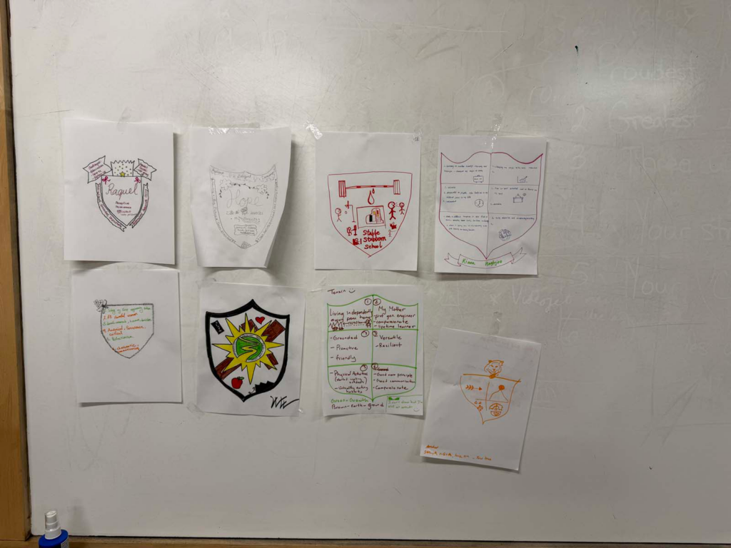One a white board, eight white pieces of paper are taped to display personalized drawings of a Coat of Arms. The papers differ from plain pencil script, to multi-colored with reds, oranges, yellows, and green.