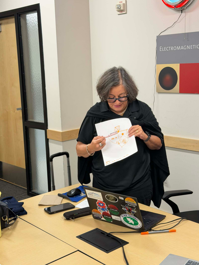 A woman stands at a conference table holding up an illustration, glancing down at the illustration pointing the different part of her illustration.