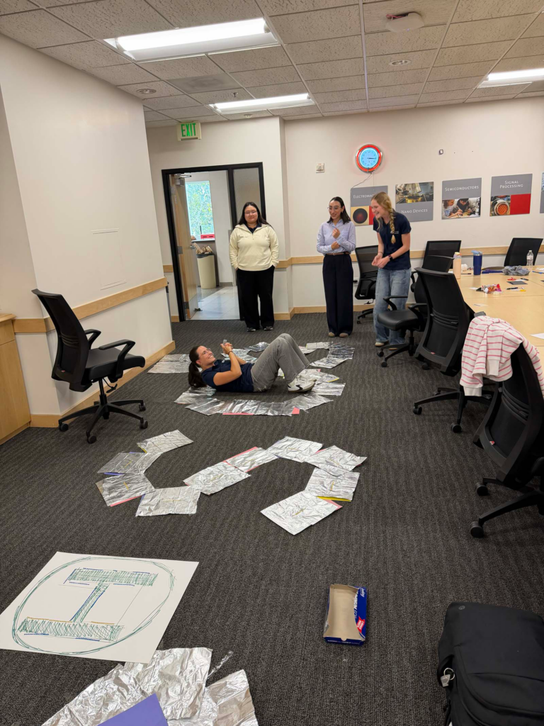 A group of four people in a University of Utah small conference room, stand on the floor with arms rearranging aluminum foil sheets in an "SOS" shape. One of the four lay on the ground in the "O" shape. A conference room table and chairs are pushed to the side of the classroom with a doorway with a red clock and engineering posters on the back wall.