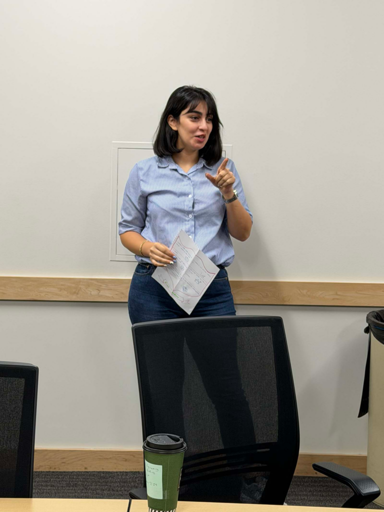 There is a woman standing in front of an off-white painted wall wearing a blue button-down shirt with jeans. She holds a personalized coat of arms, and a conference chair blocks the lower half of her legs in the center of the photo.