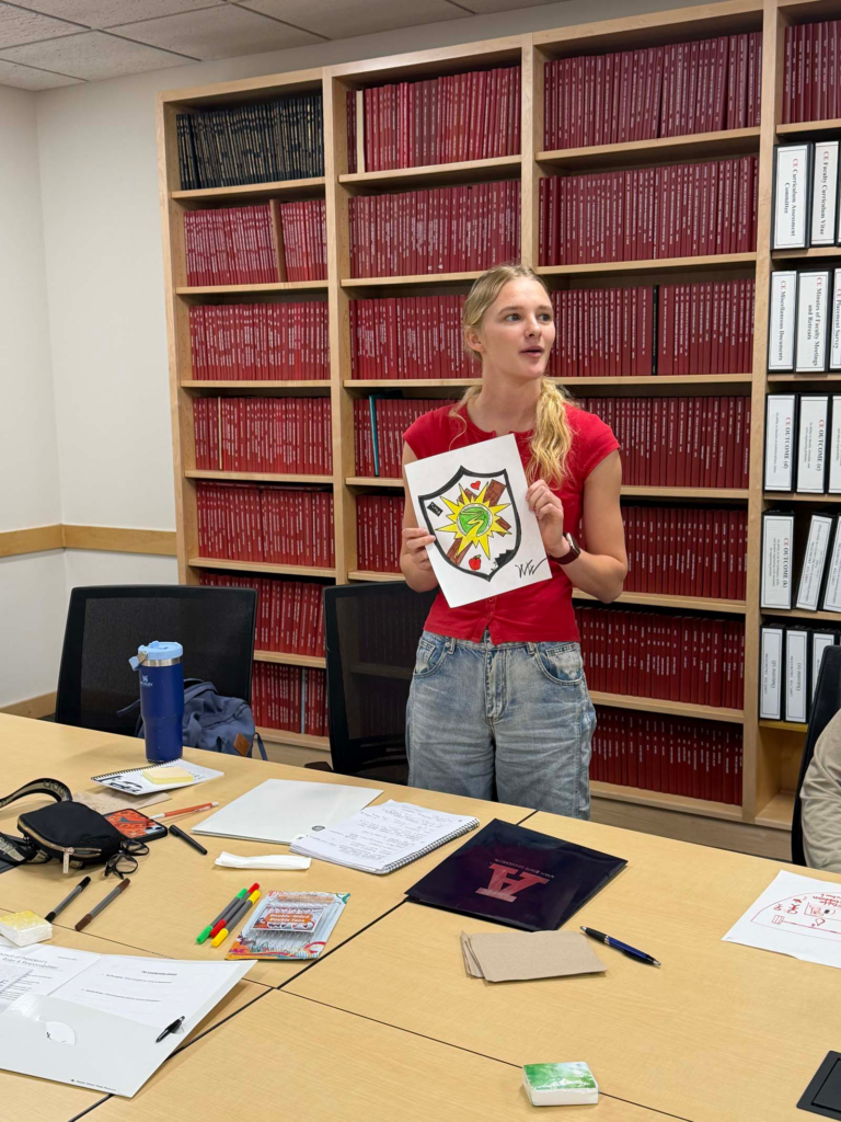 A woman wearing a red t-shirt is the centerpoint of the photo. She holds a sheet of paper that has a personalized coat of arms on display with yellow and green colors displayed on it. Behind her a built-in bookshelf holds thick red textbooks.