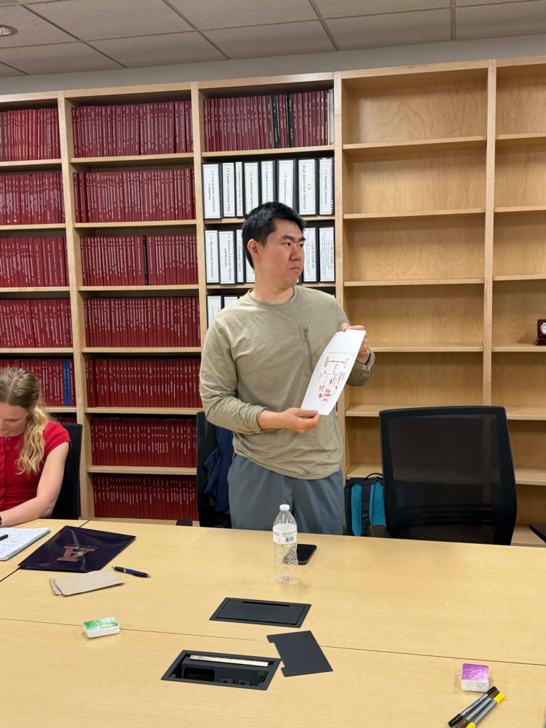 A man wearing a grey-green long-sleeved t-shirt is the centerpoint of the photo. He stands holding a sheet of paper that has a personalized coat of arms on display with red marker. Behind him there is a built-in bookshelf holds thick red textbooks and white large three ringed binders.