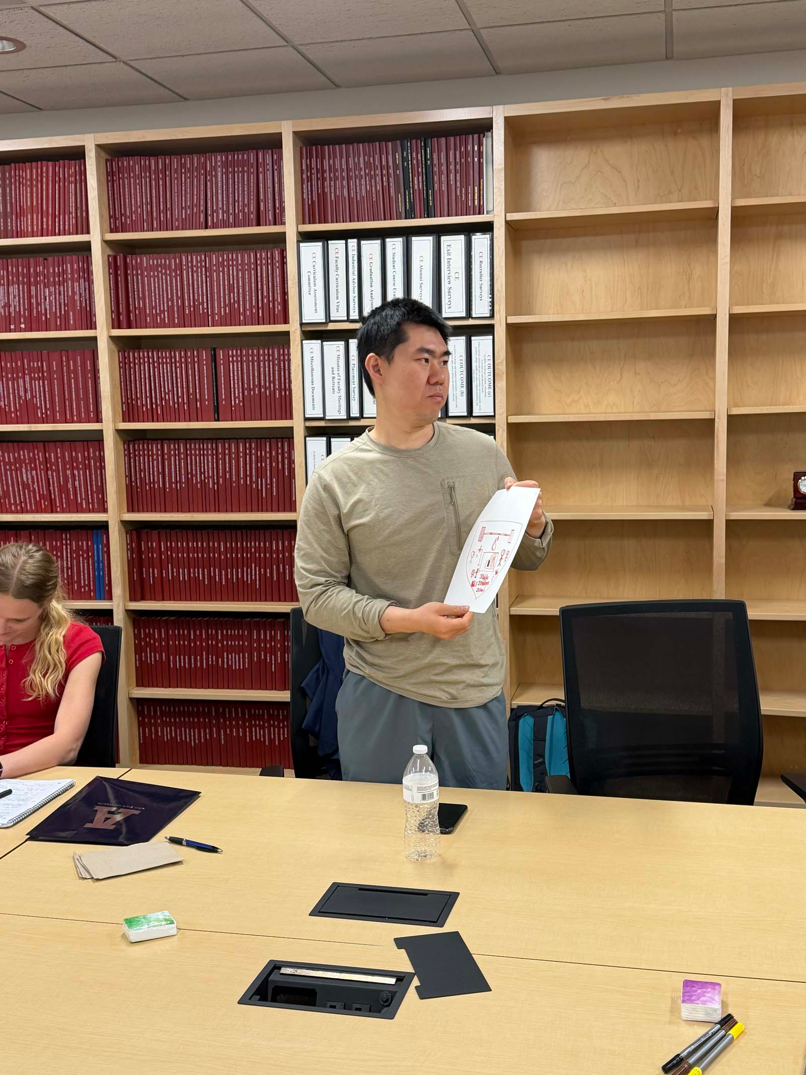A man wearing a grey-green long-sleeved t-shirt is the centerpoint of the photo. He stands holding a sheet of paper that has a personalized coat of arms on display with red marker. Behind him there is a built-in bookshelf holds thick red textbooks and white large three ringed binders.