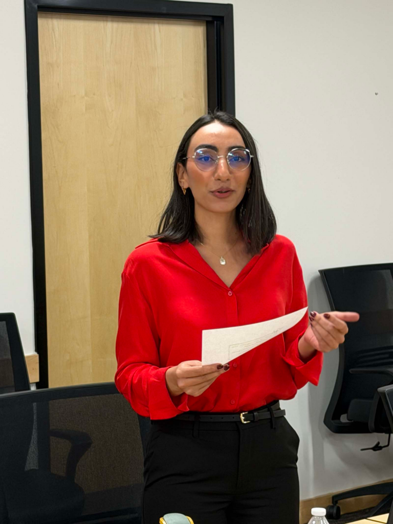 A woman wearing a red blouse is the centerpoint of the photo. She holds a sheet of paper while talking with her eyes glancing to the camera. There is a beige door and chairs behind her with an off-white color on the walls.