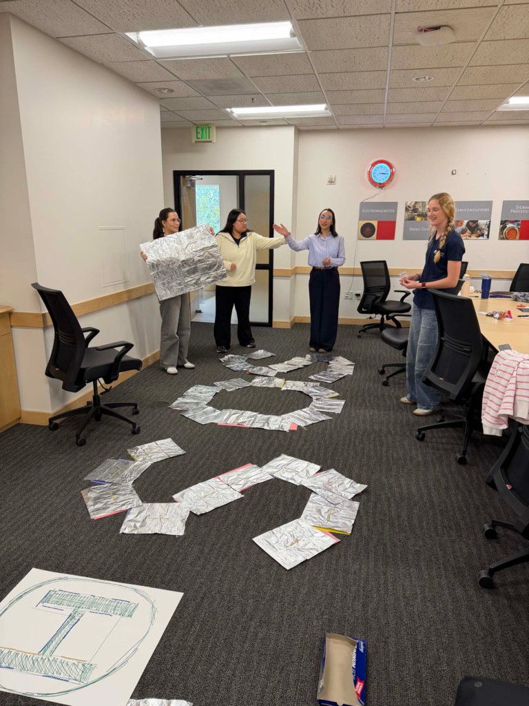 A group of four people in a University of Utah small conference room, stand on the floor with arms rearranging aluminum foil sheets in an "SOS" shape. A conference room table and chairs are pushed to the side of the classroom with a doorway with a red clock and engineering posters on the back wall.