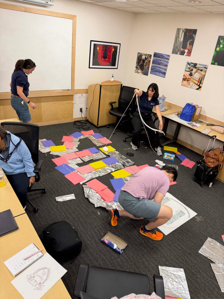 A group of four people in small conference room, stand and kneel over the floor with arms rearranging aluminum foil sheets and multicolored sheets of paper with scissors, markers, and flashlight on the ground. One woman holds a long roll of string together. A woman sits in a conference room chair next to the group of students arranging the sheets of paper and tinfoil.