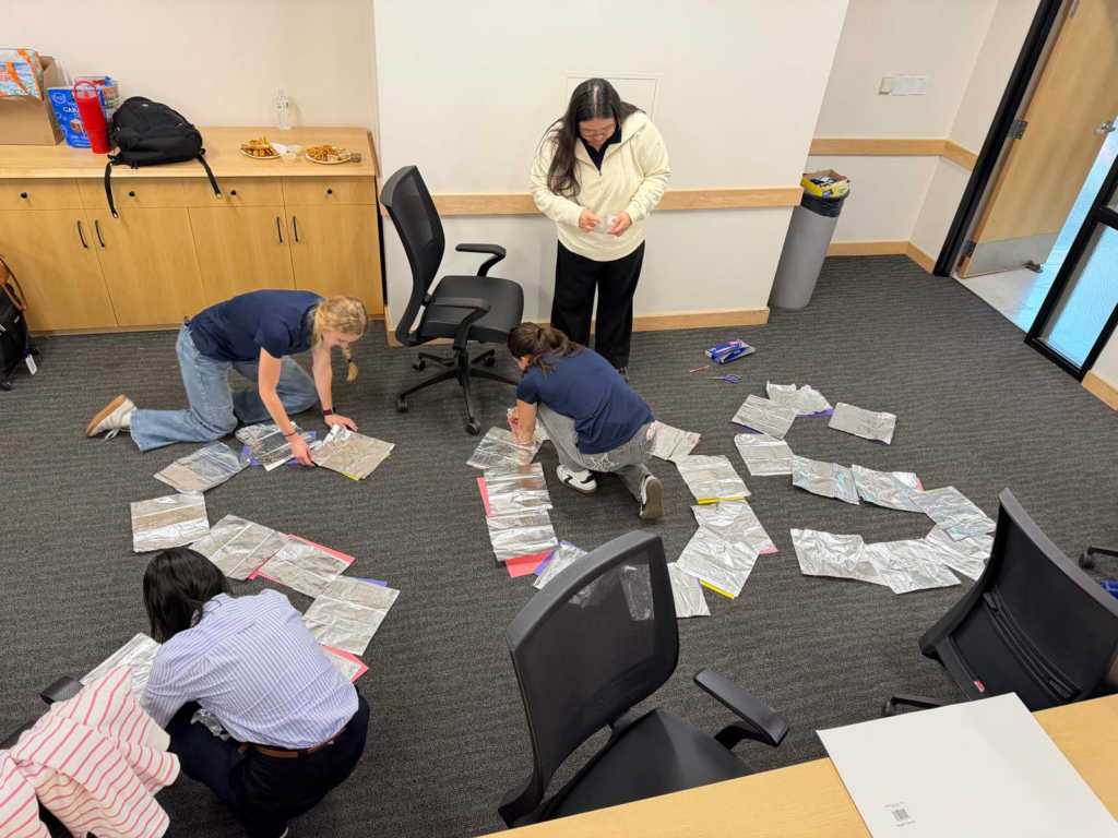 A group of four people in a University of Utah small conference room, stand and kneel on the floor with arms rearranging aluminum foil sheets and multicolored sheets of paper in an "SOS" shape. A conference room table and chairs are pushed to the side of the classroom with a doorway with a trashcan in the background, and a cabinet counter in the background holding food items and a backpack.