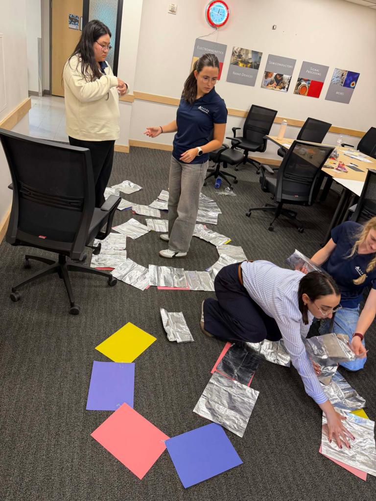 A group of four people in a University of Utah small conference room, stand and kneel on the floor with arms rearranging aluminum foil sheets and multicolored sheets of paper in an "SOS" shape. A conference room table and chairs are pushed to the side of the classroom with a doorway in the background with a red clock on the wall and engineering pictures on the wall.