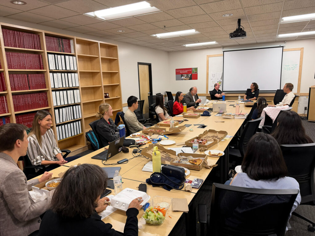 A large group of people sit around a long rectangular conference table. On the table there are pizza boxes and plates with half eaten pizza slices on them. People are actively engaging with each other across the whole table. Behind the table on the left-hand side of the photo there is a large built-in bookshelf with large amounts of books on them. Behind the head of the table is a clean white board and blank projection screen.