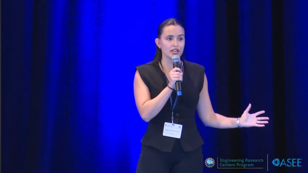A woman wearing a business suit, wearing a name tag lanyard while holding a microphone speaks with an arm outstretched. Behind the woman, a large dark curtain is in the background with blight light glowing blue behind the curtain.