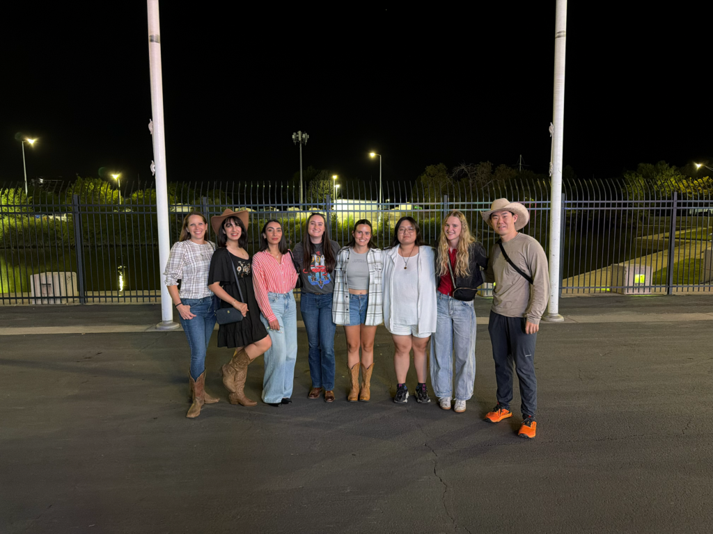 A group of eight people stand Infront of a fence with two large flag poles behind them. In the background there are multiple outdoor lamps emitting a yellow glow in the black sky. They are standing on dark grey asphalt, and four of the people are wearing cowboy boots and two are wearing cowboy hats. 