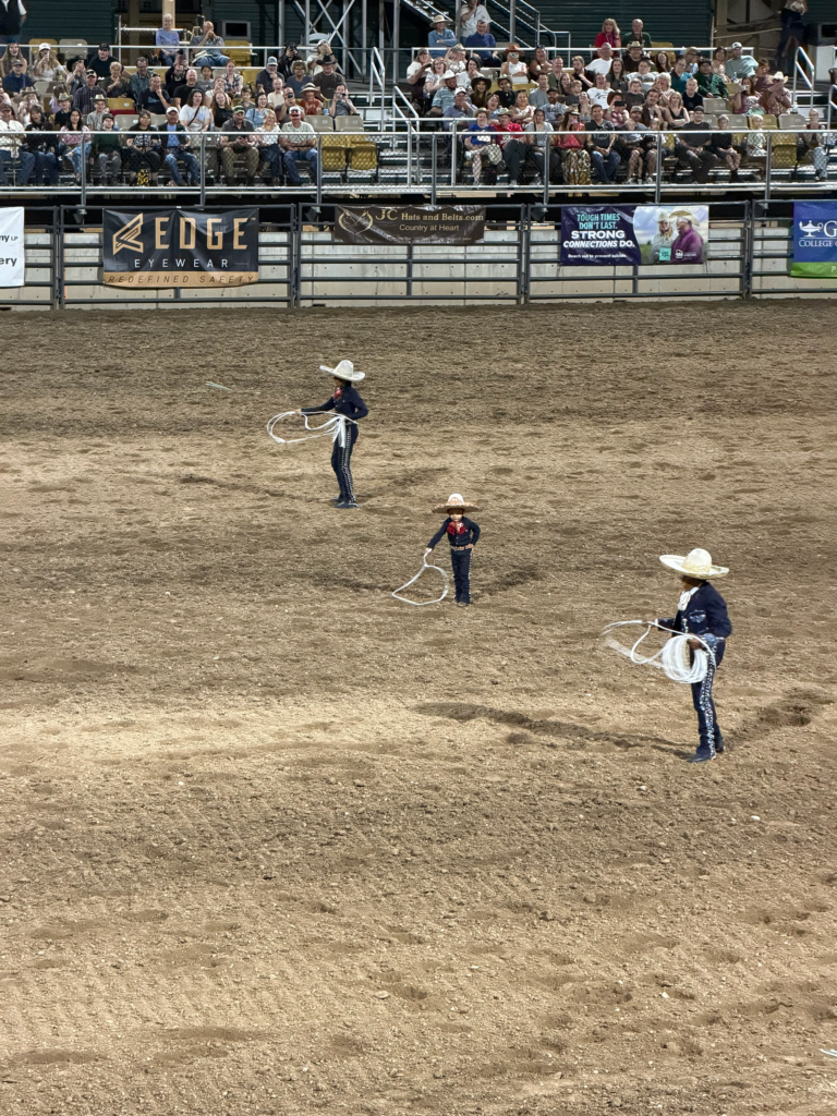 Three people stand in a diagonal two wearing traditional blue Charro outfits with lassos, standing in a sandy dirt pit. There is a riding arena fence often used in rodeos and for livestock. Banners of sponsors are on the hung up on the fence. Two people holding their lassos are swung in the air, and the shorter person in the middle of the line's lasso is pointed toward the ground. Above the sponsored lined fence, there are packed rows of people sitting in the stands. 