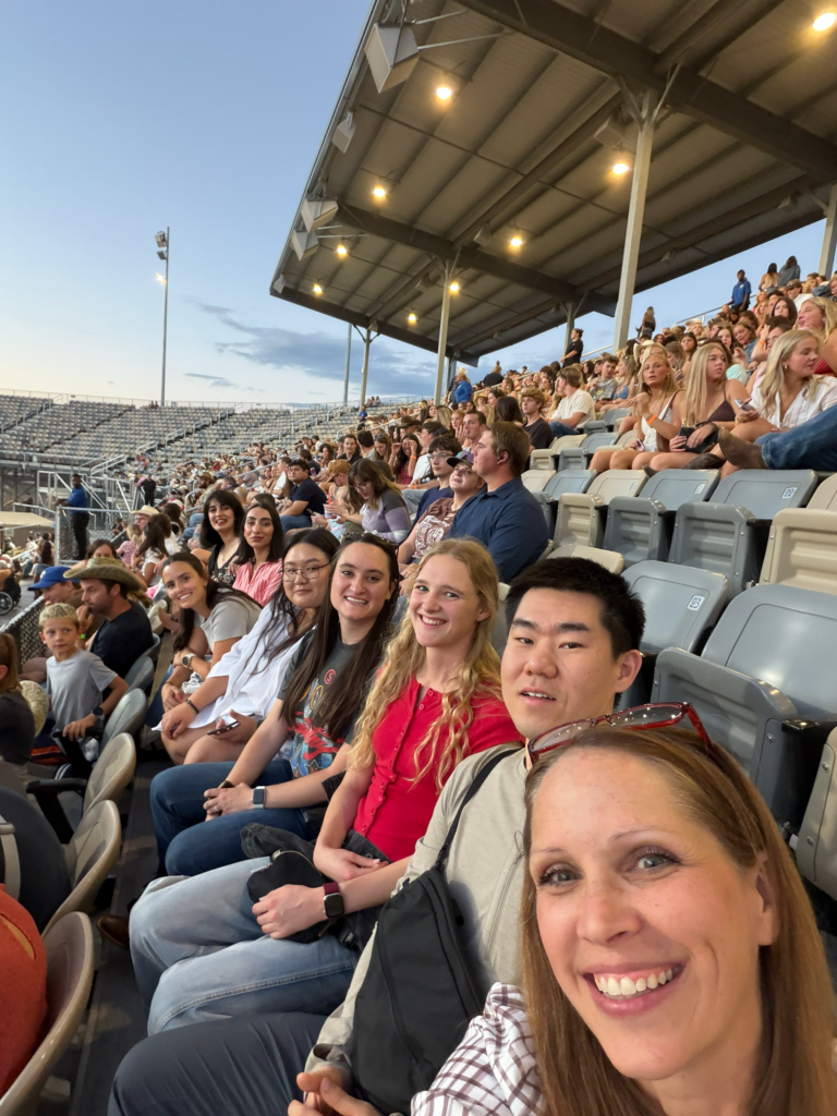 Four people sit next to one another in a large outdoor stadium with a packed crowd behind the row of people smiling toward the camera. The sky showing in the picture is an ombre from orange to a darker blue; the sun is setting. 